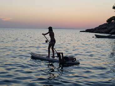 Deux personnes font du paddleboard au coucher du soleil. L'un est debout et pagaie, l'autre se détend sur la planche. Les eaux calmes et le ciel serein complètent cette scène paisible.