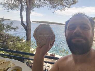 Un homme barbu sourit en tenant un morceau de pain plat au bord de la mer. On aperçoit des arbres, de l'eau bleue et une assiette de pain avec de la sauce.