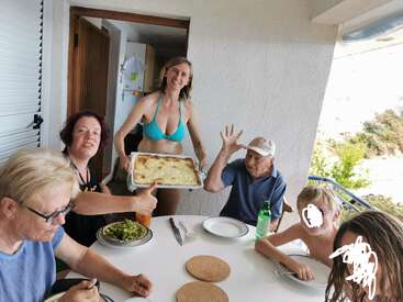 Un groupe de personnes est réuni autour d'une table sur un balcon, en train de prendre un repas ensemble. Une femme sert la nourriture, tout le monde a l'air heureux.