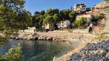 Escena costera con agua azul clara, costa rocosa y varias casas enclavadas entre pinos. La luz del sol ilumina la ladera, creando un ambiente tranquilo y acogedor.