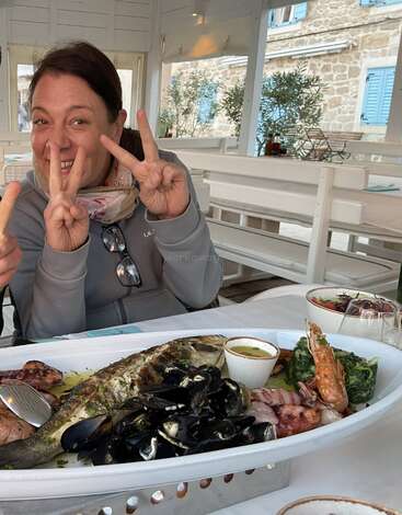 Una mujer sonríe y hace señas de paz en una mesa de un luminoso restaurante, con una gran fuente de marisco y verduras delante.