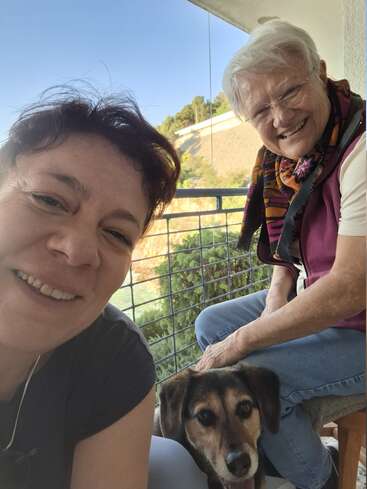 Dos mujeres, una más joven y otra más mayor, sonríen cariñosamente a la cámara sentadas al aire libre en un balcón. Un perro feliz se sienta entre ellas. Día soleado.