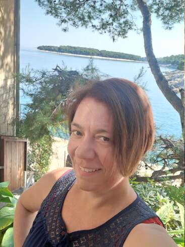 Uma mulher sorri para uma selfie ao ar livre, com vegetação exuberante, paredes de pedra, um belo mar azul e uma ilha distante ao fundo, sob um céu limpo.