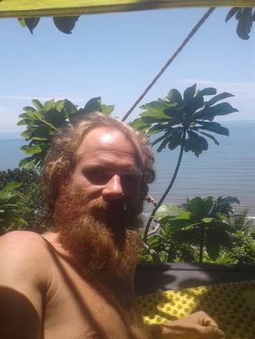 A imagem mostra um homem com barba e cabelos longos, sem camisa, em frente a um corpo d'água e uma vegetação exuberante em um dia ensolarado.