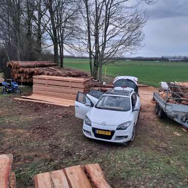 Une voiture argentée aux portes ouvertes est garée près d'un tas de planches de bois, de bûches et d'une remorque, dans une zone rurale parsemée de champs verdoyants et d'arbres.