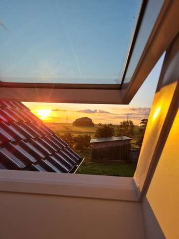 Ein atemberaubender Blick durch ein Fenster auf den Sonnenuntergang. Das Sonnenlicht durchflutet die Landschaft und beleuchtet Dächer, einen Holzschuppen, grüne Felder, ferne Bäume und einen ruhigen blauen Himmel.