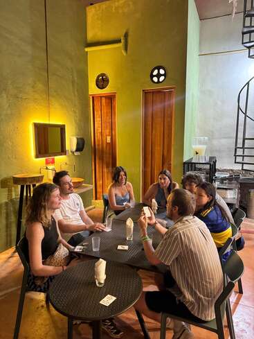 Seven people are sitting around two black tables in a cozy, warmly-lit room, engaged in conversation and playing a card game, surrounded by modern decor.