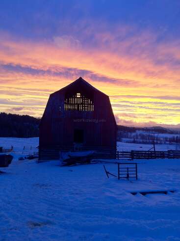 Eine rustikale Holzscheune steht in der Abenddämmerung inmitten von verschneiten Feldern und hebt sich von einem dramatischen, farbenfrohen Himmel in Orange, Rosa und Blau ab, der sich von sanften Hügeln abhebt.
