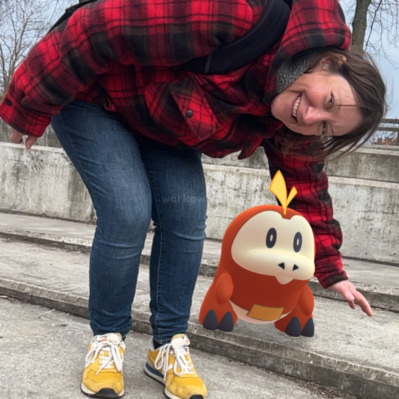 Eine Frau in einer roten Karojacke und gelben Schuhen lächelt, während sie sich draußen neben einer niedlichen, cartoonartigen orangefarbenen Pokémon-Figur namens Fuecoco auf einer Betonstufe bückt.