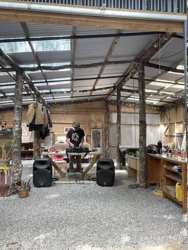 Una persona hace de DJ con su equipo bajo un cobertizo rústico de tejado abierto. Grandes altavoces, ropa colgada, plantas y varios muebles rodean el ambiente relajado y creativo.