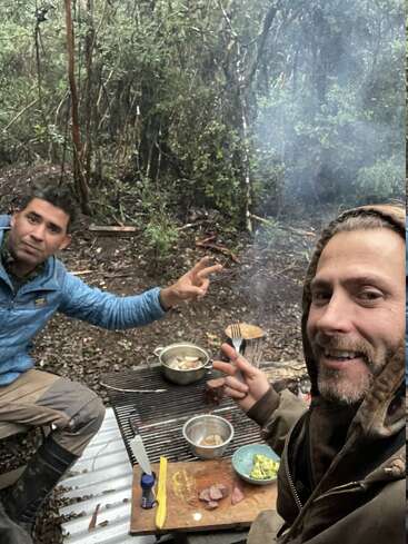 Dos hombres sentados al aire libre en un bosque, sonríen y hacen signos de paz mientras cocinan alimentos en un fuego abierto. Se ven utensilios de cocina, comida y humo.