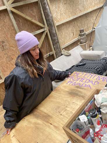 Una mujer con gorro morado y chaqueta negra pinta un cartel de madera en el que se lee "No Entrar Propiedad Privada" dentro de una rústica estructura de madera inacabada con material artístico cerca.