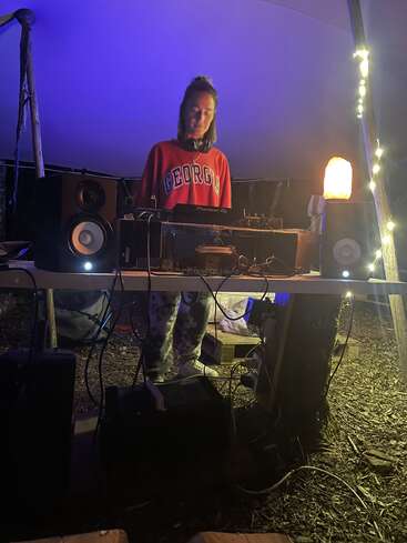 Una persona con una sudadera roja de "Georgia" ejerce de DJ al aire libre por la noche, bajo una carpa, rodeada de altavoces, luces, cables y una lámpara incandescente, creando ambiente.