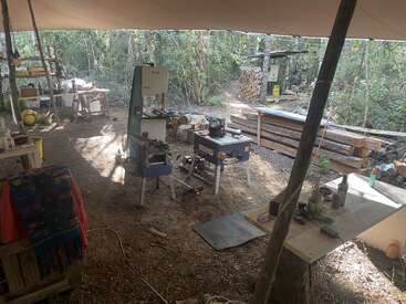 Un taller de carpintería al aire libre en el bosque, cubierto por una lona, con varias herramientas, equipos, bancos, pilas de madera y mesas rodeadas de árboles y luz solar.