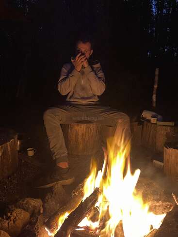 Una persona se sienta en un tocón de madera junto a una hoguera por la noche, calentándose mientras bebe de una taza, rodeada de árboles y asientos rústicos.
