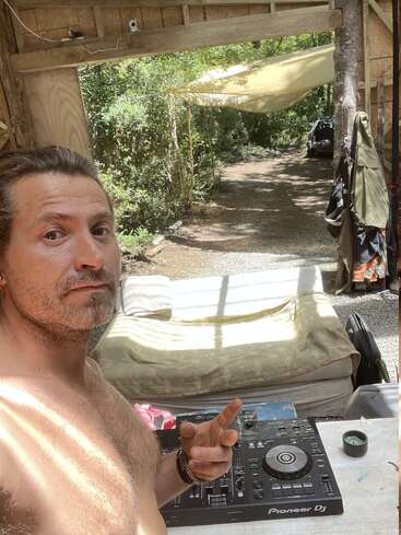 Un hombre sin camiseta se hace un selfie en un entorno rústico al aire libre con frondosos árboles. Muestra un signo de la paz cerca de un controlador de DJ Pioneer sobre una mesa.