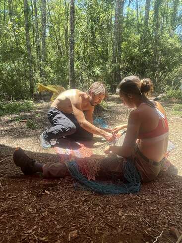 Dos personas sentadas en el suelo en un bosque iluminado por el sol, trabajando juntas en una colorida red. La luz del sol se filtra entre los árboles, creando una escena de tranquilidad y colaboración al aire libre.