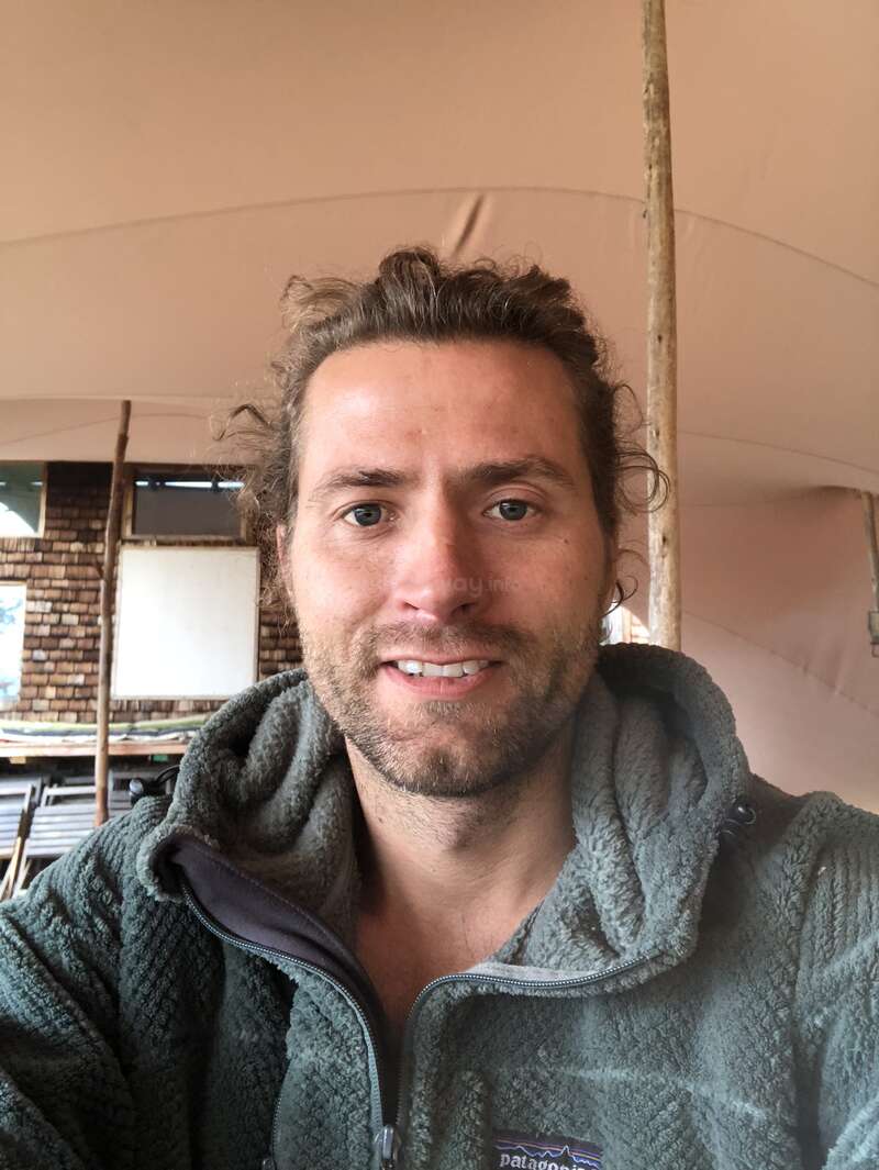 La imagen muestra a un hombre con pelo oscuro y barba, vestido con una chaqueta gris, de pie delante de un edificio con tejado blanco y paredes de ladrillo.