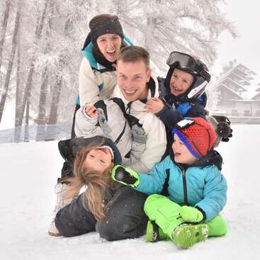Eine fröhliche fünfköpfige Familie spielt zusammen im Schnee, eingepackt in Winterkleidung, lächelnd und lachend, umgeben von frostigen Bäumen und einer verschneiten Winterlandschaft.