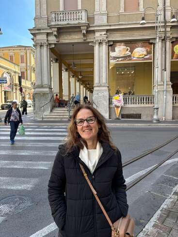 A woman in a black jacket stands smiling on a crosswalk, with an elegant building, street, and people in the background, on a bright day.