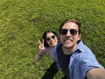The image shows a man and woman taking a selfie in a grassy field, with the woman making a peace sign and both wearing sunglasses.