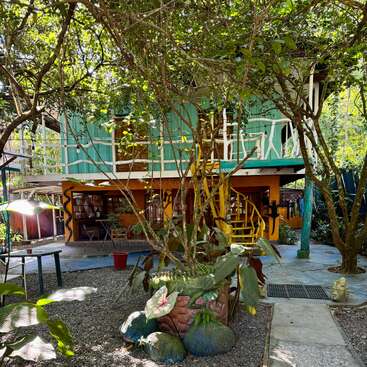 The image depicts a vibrant two-story building with a balcony, surrounded by lush trees and plants, set against a serene outdoor backdrop.