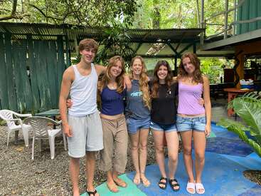 Esta imagem mostra cinco pessoas juntas ao ar livre, sorrindo. Elas estão vestidas casualmente, descalças ou de sandálias, cercadas por vegetação e móveis de pátio, parecendo felizes e relaxadas.