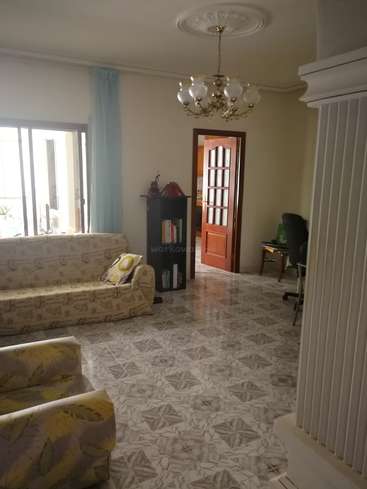 La imagen muestra un salón con un sofá, un sillón, una estantería y una lámpara de araña, con paredes blancas, suelo de baldosas y una puerta que da a otra habitación.