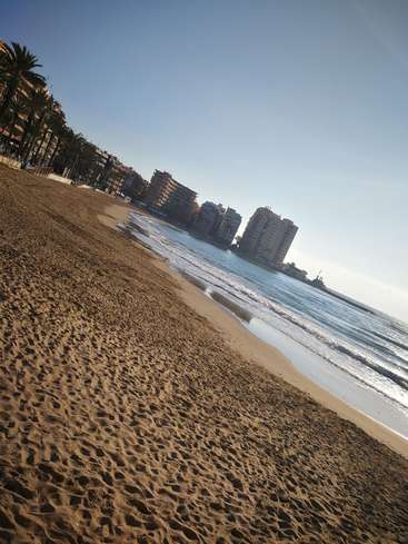 La imagen representa una serena escena playera con una orilla de arena, suaves olas y una hilera de edificios al fondo, sobre un cielo azul despejado.