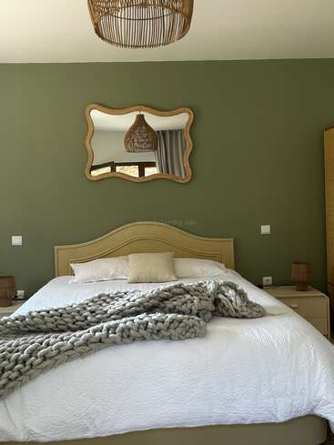 Cette chambre chaleureuse se distingue par un mur d'accent vert, un miroir au cadre ondulé, un lit beige recouvert d'une couverture en grosse maille, ainsi que des éléments décoratifs en osier au plafond et sur les lampes.