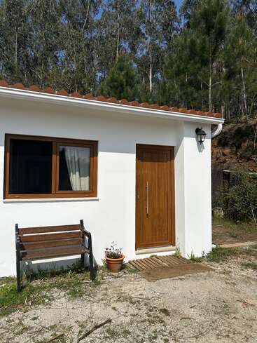 Une petite maison blanche, avec une porte et une fenêtre en bois, se trouve au milieu d'un bosquet. À l'extérieur, on trouve un banc, une plante en pot et une lanterne.