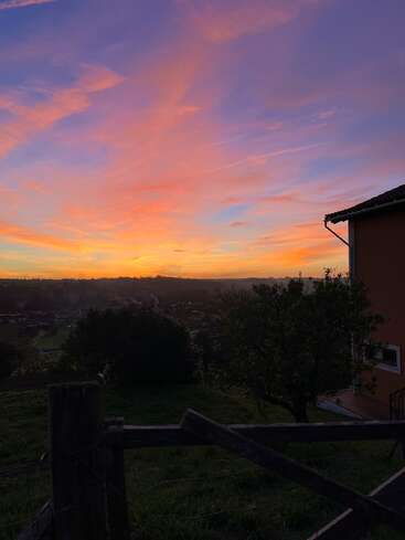 Um pôr do sol vibrante pinta o céu com tons de laranja, rosa e roxo. Árvores em silhueta, uma cerca de madeira e uma casa completam essa cena tranquila do campo.