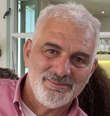 Um cara de meia-idade, com cabelo branco curto e encaracolado e barba branca bem aparada, sorri calorosamente. Ele usa uma camisa xadrez vermelha dentro de uma sala bem iluminada.