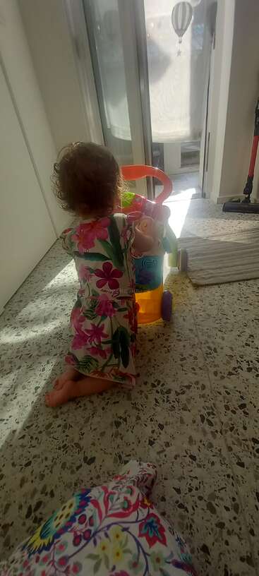 Uma criança pequena, vestida com um vestido florido, está ajoelhada no chão de terrazzo, brincando com um carrinho colorido perto de uma porta de vidro. A luz do sol ilumina o cômodo.