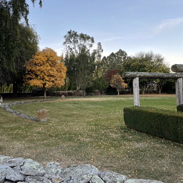 The image depicts a serene grassy area with a stone wall, trees, and a wooden structure, set against a blue sky with white clouds.