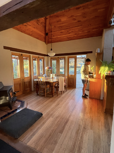 The image depicts a warm and inviting room with a wooden floor, a dining table, and a man standing near a cabinet, surrounded by natural light and a cozy atmosphere.