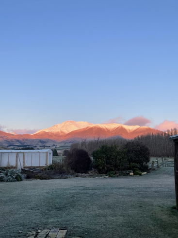 The image depicts a serene landscape featuring a snow-capped mountain range, a small white building, and a lush green field, set against a clear blue sky.