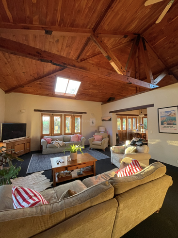 The image depicts a cozy living room with a vaulted wood ceiling, featuring a tan couch, coffee table, and two beige sofas, surrounded by windows and natural light.