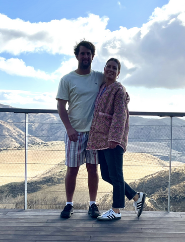 The image depicts a man and woman standing on a deck, with a mountainous landscape in the background, under a partly cloudy sky.