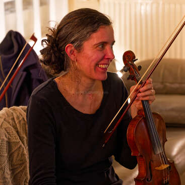 Uma mulher sorridente está sentada dentro de casa, segurando um violino e um arco. Ela veste uma roupa preta, tem cabelos cacheados presos para trás e parece relaxada e feliz enquanto está sentada.