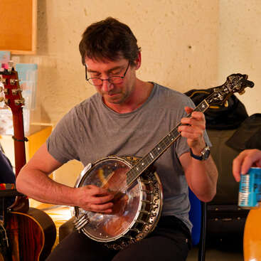 Um homem está sentado numa cadeira azul, tocando banjo com concentração. Ele usa óculos e uma camisa cinza, cercado por outros instrumentos musicais dentro de casa.