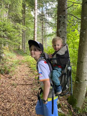 Un niño pequeño camina por un frondoso bosque verde, llevando a un bebé sonriente en una mochila portabebés. La luz del sol se filtra a través de los altos árboles, creando una atmósfera de tranquilidad.