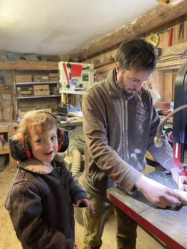 En un acogedor taller, un niño sonriente con orejeras está de pie junto a un adulto que maneja una máquina para trabajar la madera. Las herramientas y las estanterías de madera llenan el fondo con un ambiente cálido.
