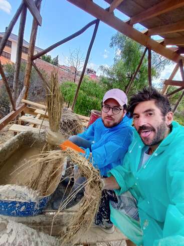 Deux hommes vêtus d'imperméables colorés travaillent avec de la boue et de la paille, probablement pour fabriquer des matériaux d'éco-construction, en souriant sous une structure en bois par une journée ensoleillée.