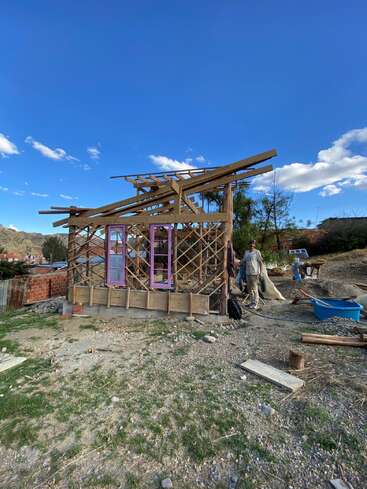 Une petite maison en bois en construction avec des poutres apparentes et des cadres de fenêtres violets, située à l'extérieur par une journée ensoleillée. Une personne travaille à proximité.