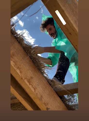 Un homme vêtu d'un imperméable vert travaille sur un toit de chaume, manipulant de la paille entre des poutres en bois, sous un ciel bleu et quelques nuages visibles.