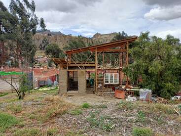 Ein teilweise gebautes Haus mit Holzrahmen und einigen Wänden steht zwischen Bäumen und Gras. Im Hintergrund erscheinen Berge und Häuser unter bewölktem Himmel.