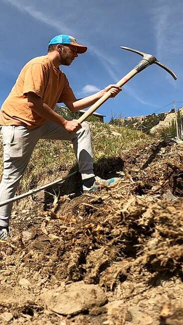 Ein Mann mit blauer Mütze und orangefarbenem Hemd benutzt an einem sonnigen Tag eine Spitzhacke und gräbt entschlossen in den felsigen Boden eines Hügels.