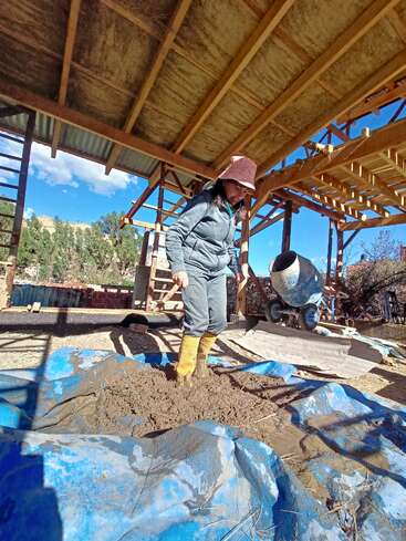 Eine Person mit gelben Stiefeln mischt Schlamm auf einer blauen Plane, wahrscheinlich für den Bau, unter einem hölzernen Dachstuhl, im Freien mit klarem Himmel und natürlicher Umgebung.