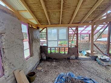 Dieses Bild zeigt das Innere eines im Bau befindlichen Hauses mit freiliegenden Holzbalken, unfertigen Wänden, Fenstern, Stein und auf dem Boden verstreuten Baumaterialien.